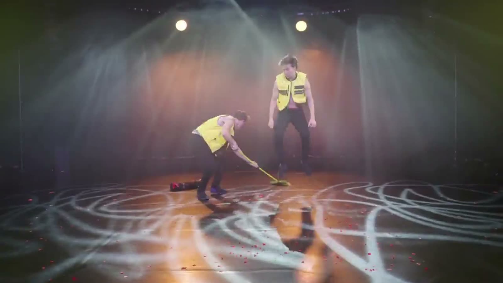 Jim & John in yellow safety vests on an empty stage under spotlights, one sweeping the floor with a yellow broom