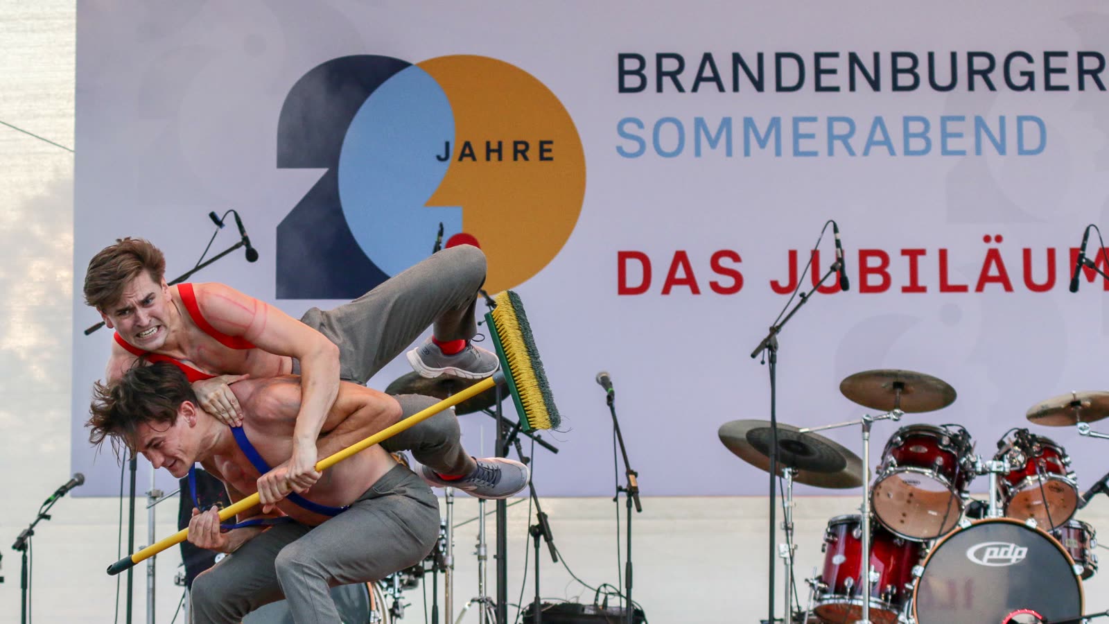 Jim & John performing their Bühnenfeger act on the open-air stage of the Brandenburger Sommerabend 20th anniversary event, the event banner and a drum kit behind them