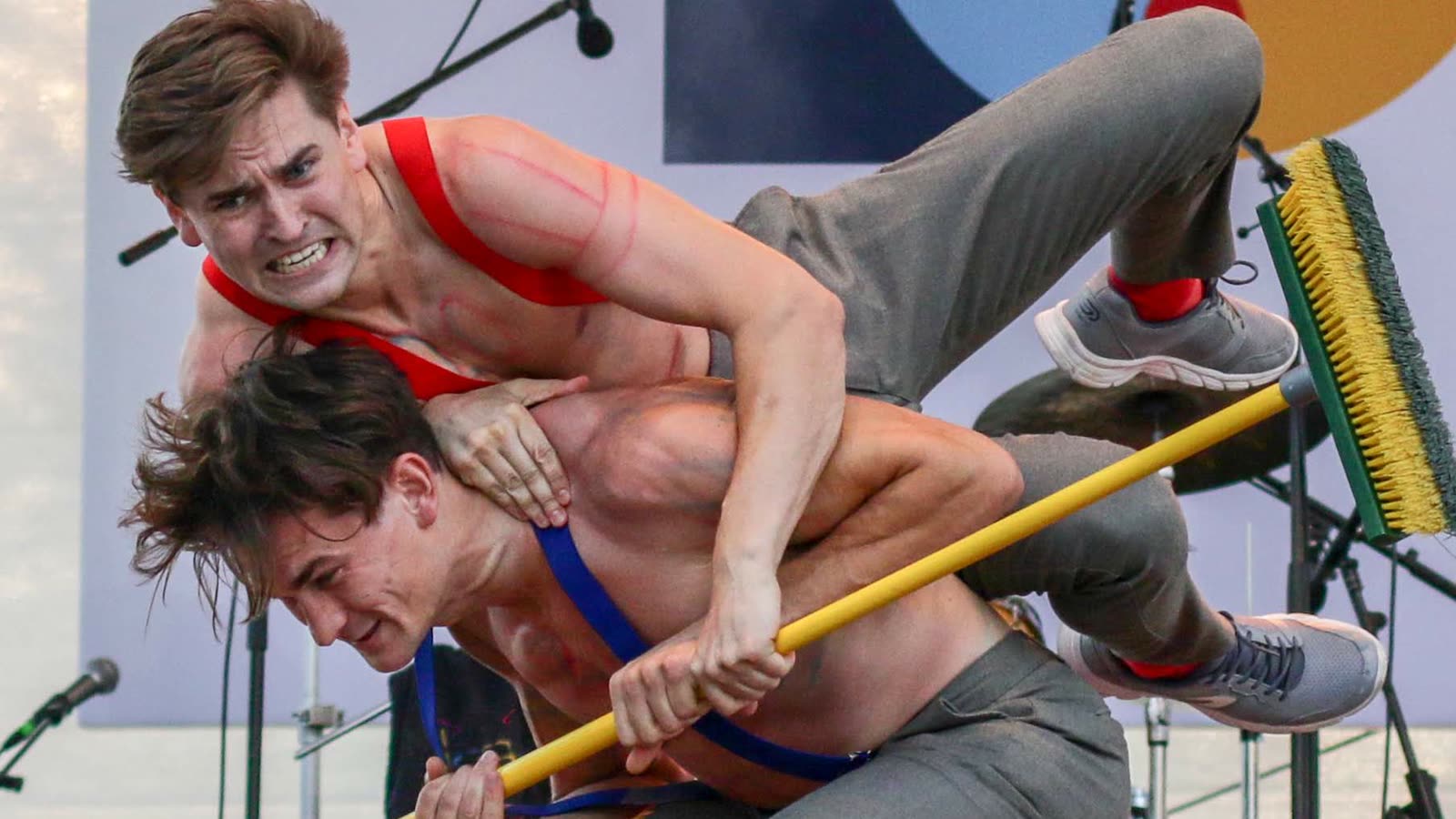 Jim and John Förster in a partner acrobatics figure on stage: the upper brother lies horizontally across his brother's shoulders, both holding a yellow broom handle