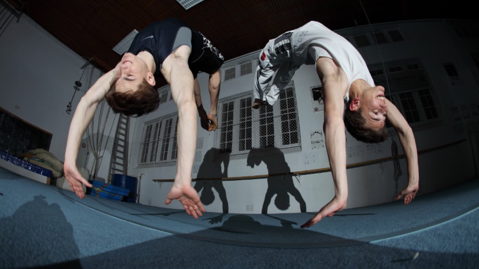 Both brothers mid-backflip upside down in the training hall, their shadows on the wall behind them